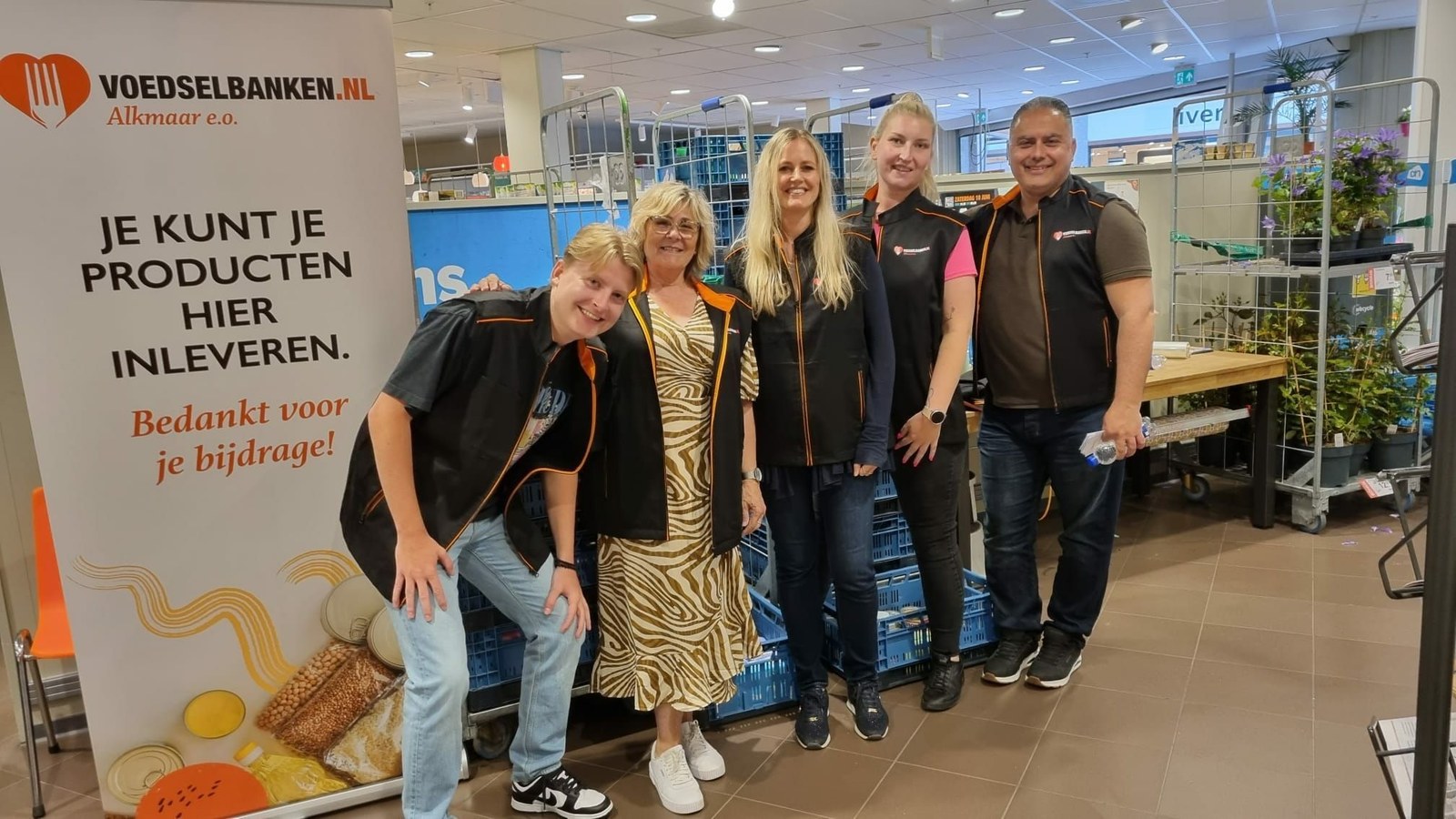 Vijf personen poseren voor een banner van Voedselbank Alkmaar in een magazijn met voedselbakken en plantjes op de achtergrond.